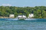 A catamaran deck boat built by Splendor Boats driving across still blue water with a forest and two large houses behind it.