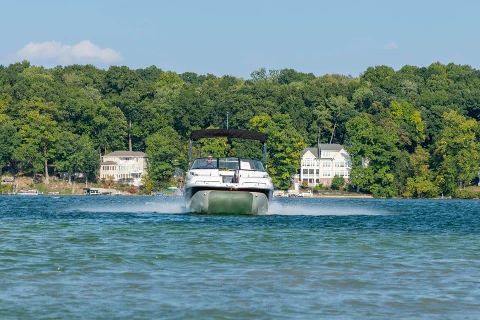 A catamaran deck boat built by Splendor Boats driving across still blue water with a forest and two large houses behind it.